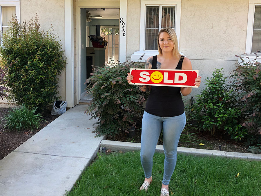 Woman standing in front of her home holding a sign that says sold with a happy face as the O.