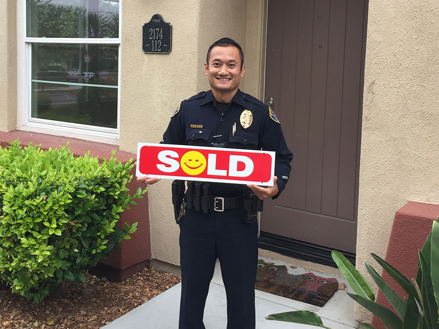 Man standing in front of a home, wearing his police uniform and holding a sign that says sold with the O as a happy face.